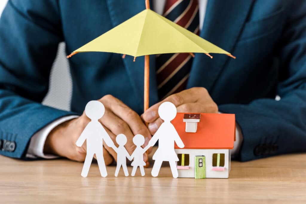 Sylwetka mężczyzny, który trzyma mini parasol nad mini domem i wycinanką rodziny, który symbolizuje ochronę jaką daje zakup ubezpieczenia.